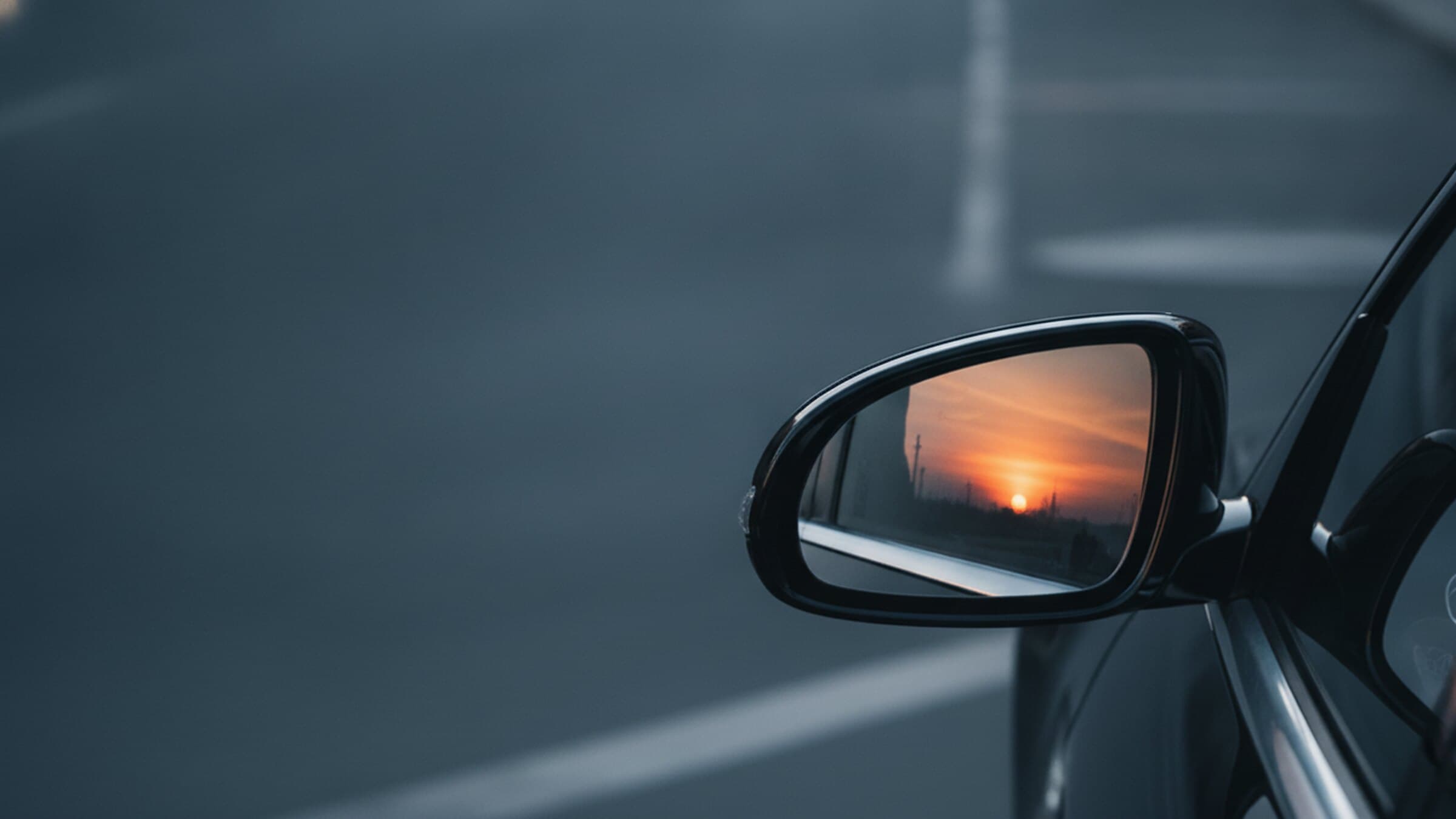 Außenspiegel eines Fahrzeugs, im Spiegelglas eine tiefstehende Sonne über einer flachen Landschaft mit Strommasten.