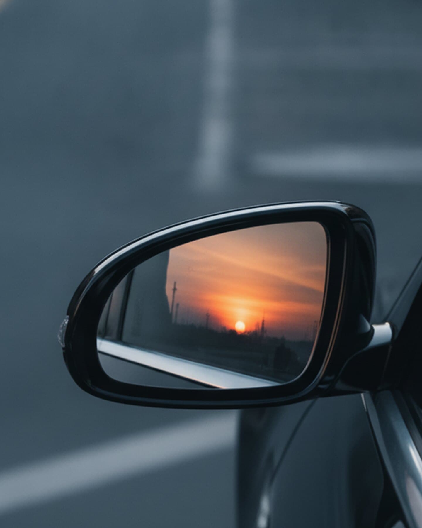 Außenspiegel eines Fahrzeugs, im Spiegelglas eine tiefstehende Sonne über einer flachen Landschaft mit Strommasten.